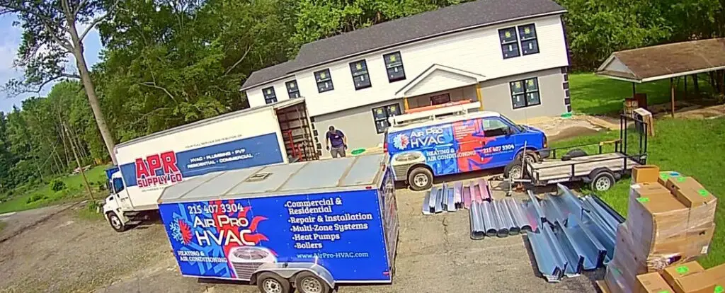 Two workers stand near HVAC company vehicles and equipment outside a two-story house, where ductwork and supplies are arranged on the driveway, ready for servicing air conditioning systems or commercial air con repairs near me.