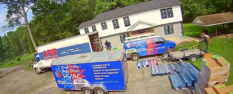 Two workers stand near HVAC company vehicles and equipment outside a two-story house, where ductwork and supplies are arranged on the driveway, ready for servicing air conditioning systems or commercial air con repairs near me.
