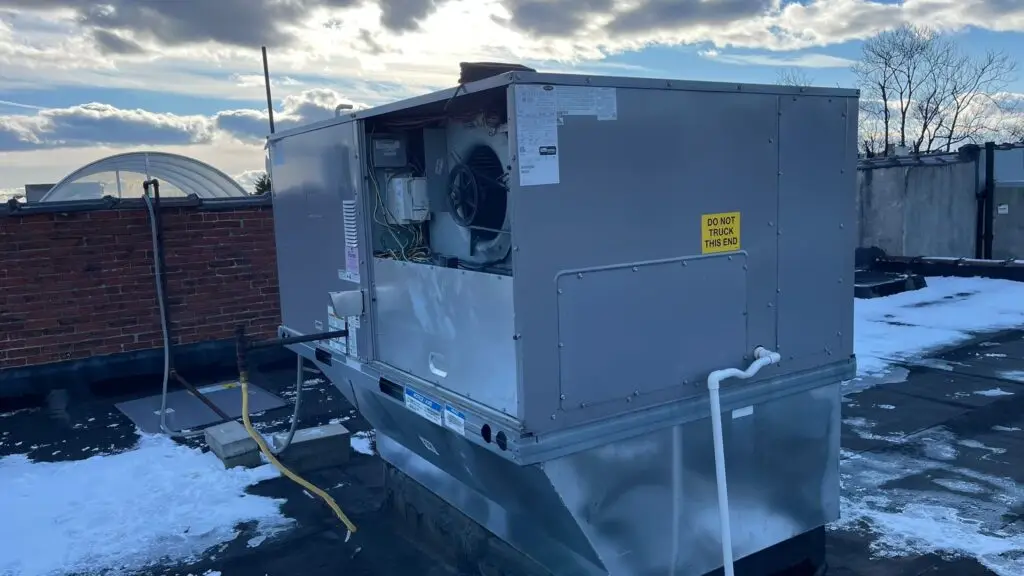 A large rooftop HVAC unit with an open panel sits on a snowy building roof under a partly cloudy sky, awaiting commercial air con repairs by an expert air conditioning repairman near me.