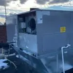 A large rooftop HVAC unit with an open panel sits on a snowy building roof under a partly cloudy sky, awaiting commercial air con repairs by an expert air conditioning repairman near me.