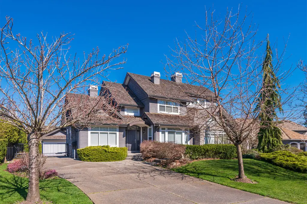 Aston, PA Two-story suburban house with gray siding, multiple chimneys, and a large driveway—ideal for servicing air conditioning—surrounded by lawn, shrubs, and leafless trees under a clear blue sky.
