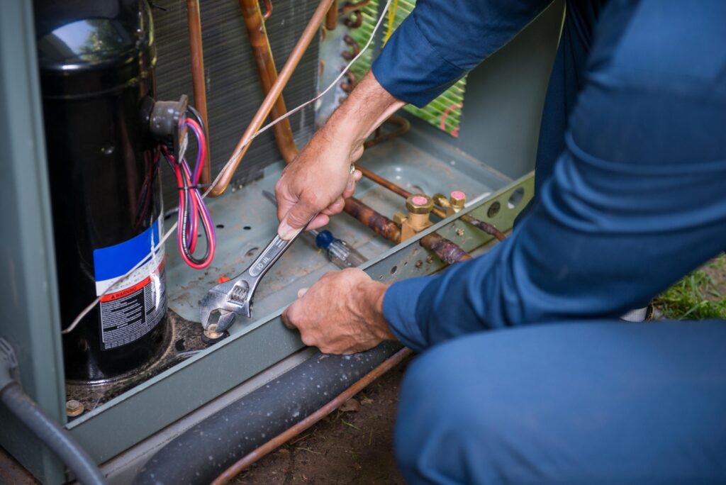 An HVAC technician uses a wrench to work on the copper refrigerant lines and components of an outdoor air conditioning condenser unit.