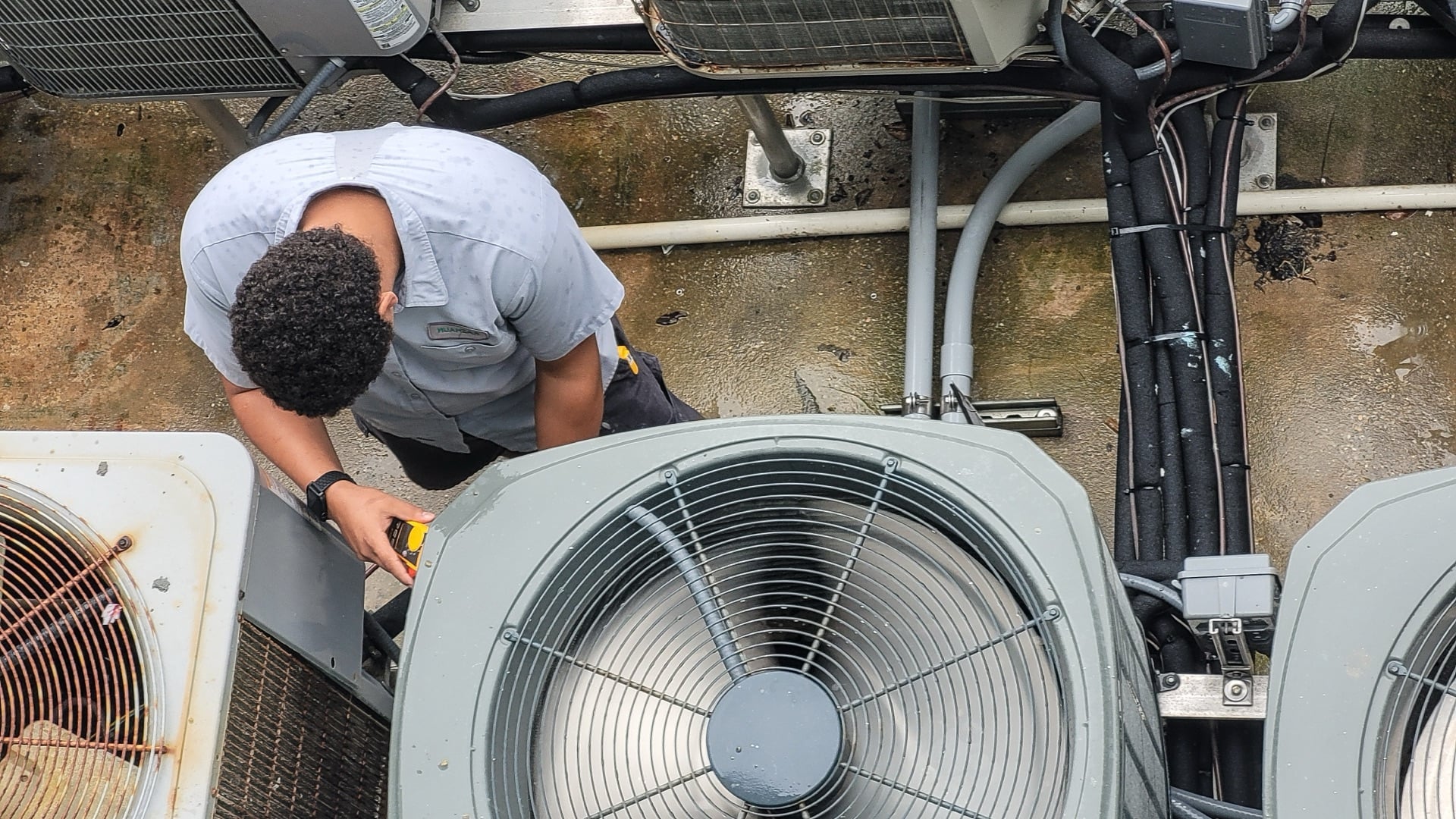 A technician in a gray uniform checks an outdoor air conditioning unit with a tool, surrounded by other HVAC units and cables—perfect for your landing page header image.