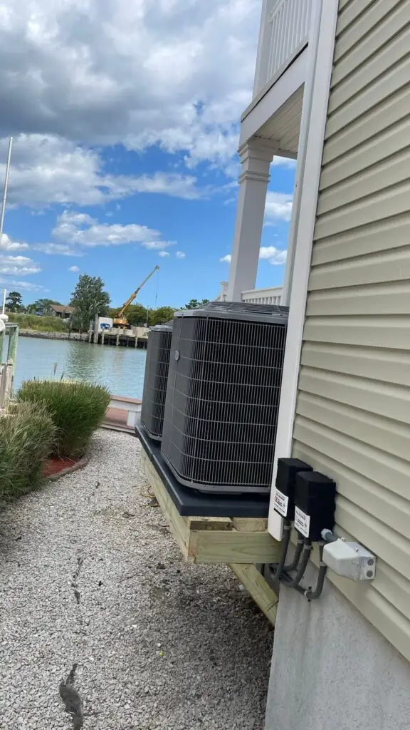 Two large air conditioning units are installed on a platform next to a house by the water, with an air conditioning repairman working nearby and construction visible across the canal.