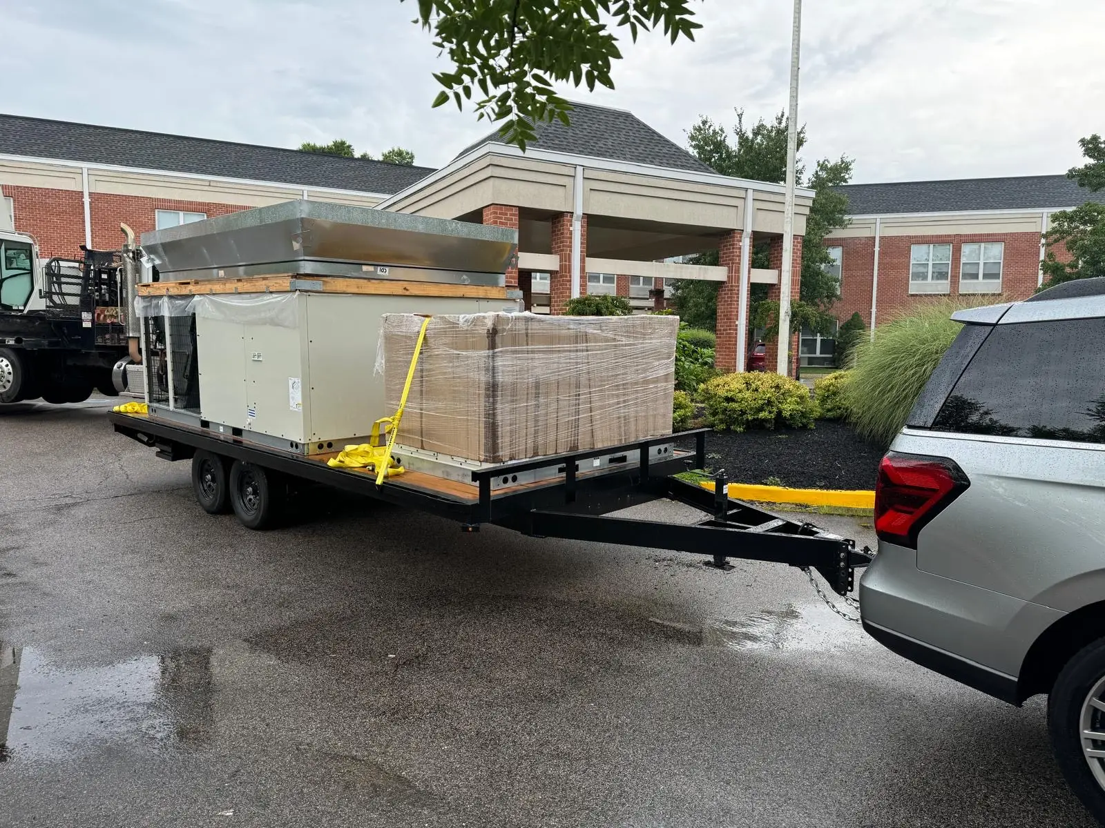 A trailer hitched to an SUV carries a large HVAC unit and a pallet of boxed goods, secured with yellow straps, parked outside a brick building for air conditioner repairing service.