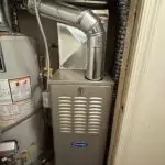 A gas furnace unit with a Carrier logo is installed next to a hot water heater in a utility closet, featuring connected vent pipes and visible wiring—ideal for servicing air conditioning or air conditioner repairing service needs.