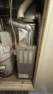 A gas furnace unit with a Carrier logo is installed next to a hot water heater in a utility closet, featuring connected vent pipes and visible wiring—ideal for servicing air conditioning or air conditioner repairing service needs.