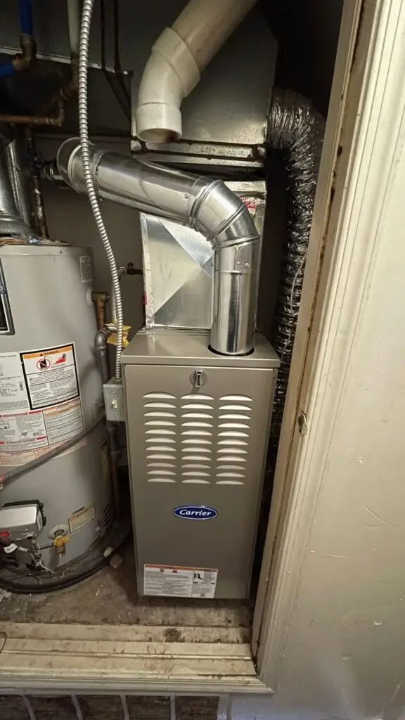 A Carrier high-efficiency gas furnace installed in a utility closet next to a water heater, with silver ductwork and a flexible exhaust pipe connected at the top.