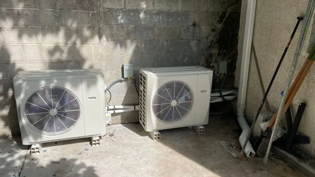 Two outdoor air conditioning units are installed on a concrete surface against a cinder block wall, with pipes, electrical boxes, and garden tools nearby—ideal for those searching "servicing air conditioning near me" or needing commercial air con repairs.