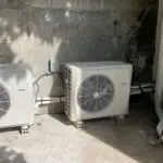 Two outdoor air conditioning units are installed on a concrete surface against a cinder block wall, with pipes, electrical boxes, and garden tools nearby—ideal for those searching "servicing air conditioning near me" or needing commercial air con repairs.