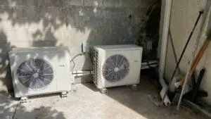 Two outdoor air conditioning units are installed on a concrete surface against a cinder block wall, with pipes, electrical boxes, and garden tools nearby—ideal for those searching "servicing air conditioning near me" or needing commercial air con repairs.