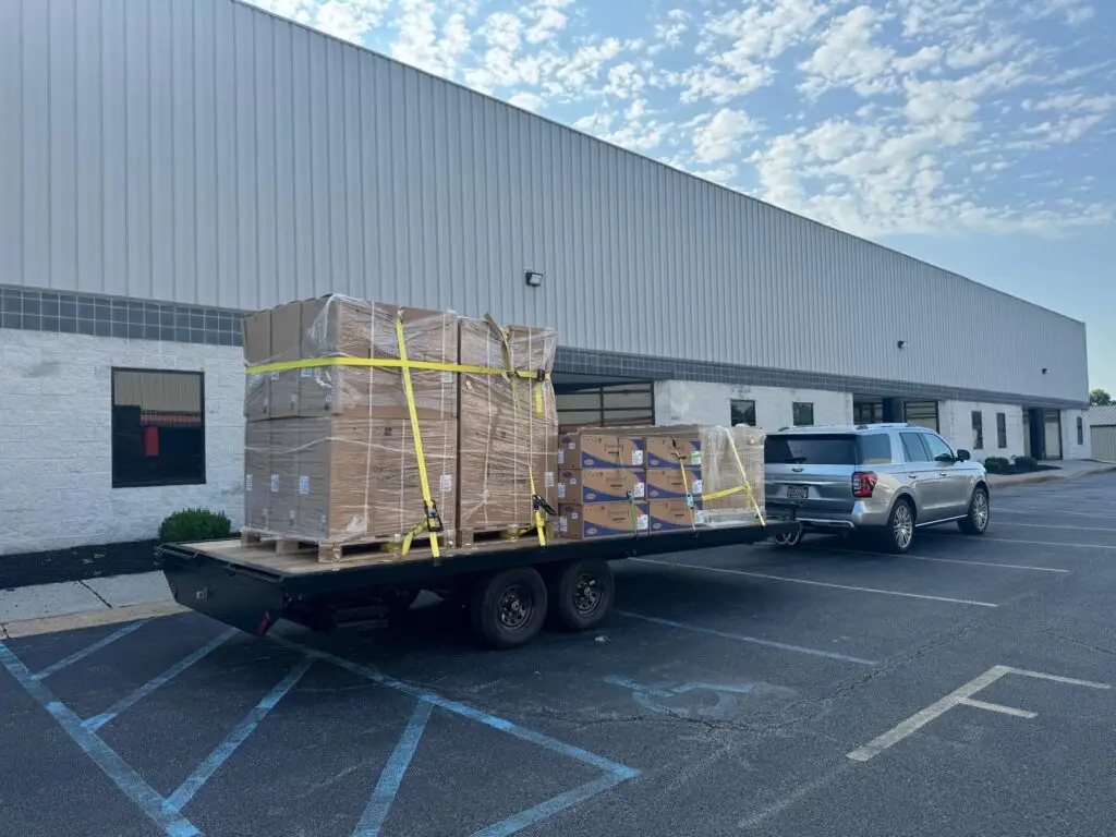 A silver SUV, possibly belonging to an air conditioning repairman, tows a flatbed trailer loaded with large, shrink-wrapped pallets and boxes outside a warehouse building.