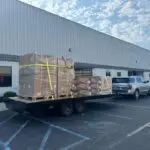 A silver SUV, possibly belonging to an air conditioning repairman, tows a flatbed trailer loaded with large, shrink-wrapped pallets and boxes outside a warehouse building.